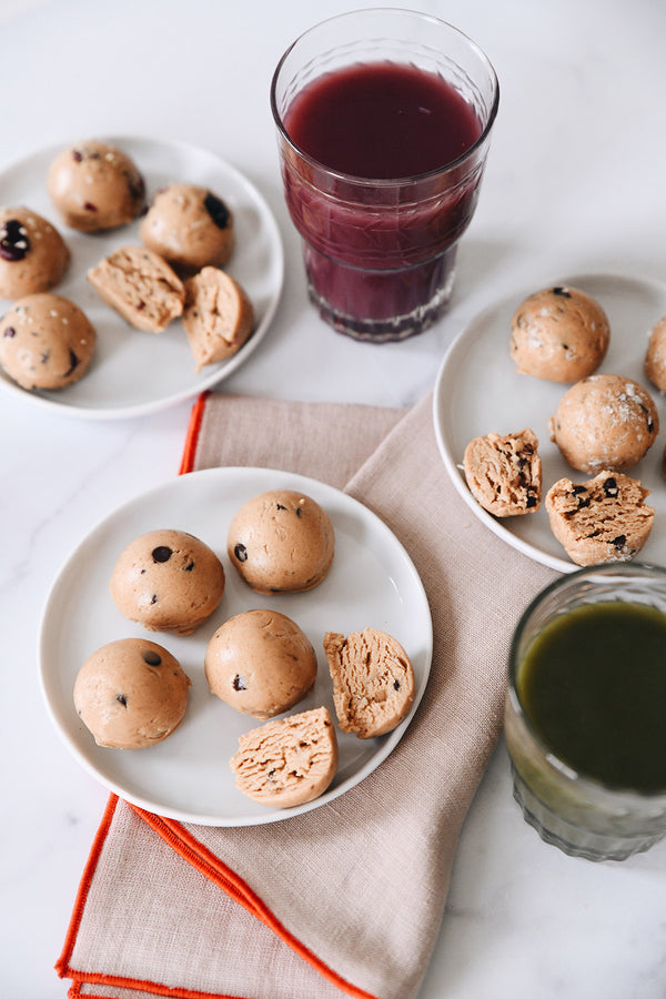 Protein bites arranged on three different plates on top of a red trimmed tea towel with glasses of green and red juice nearby.