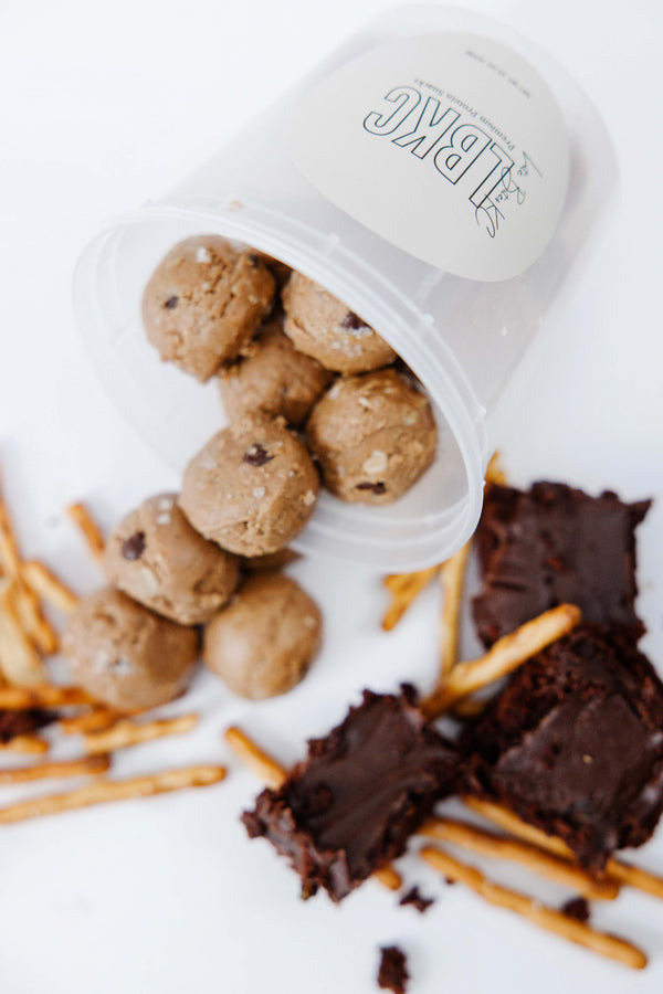 Chocolate brownie batter protein bites spilling out of container with pretzels and brownies surrounding it